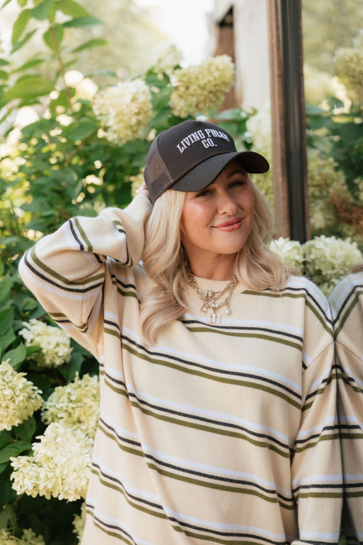 A woman wearing a cream and olive green striped knit sweater and a dark brown baseball cap with 'LIVING FULLY' embroidered on it, posing outdoors in front of hydrangea bushes.