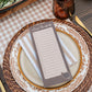 A close-up of a styled table setting featuring a rectangular notepad with lined writing space and the heading 'Living thankfully' placed on top of a place setting. The notepad is layered over a striped napkin, a decorative plate, a woven placemat, and a checkered tablecloth, with a glass, lemon slices, and a wooden serving board in the background.