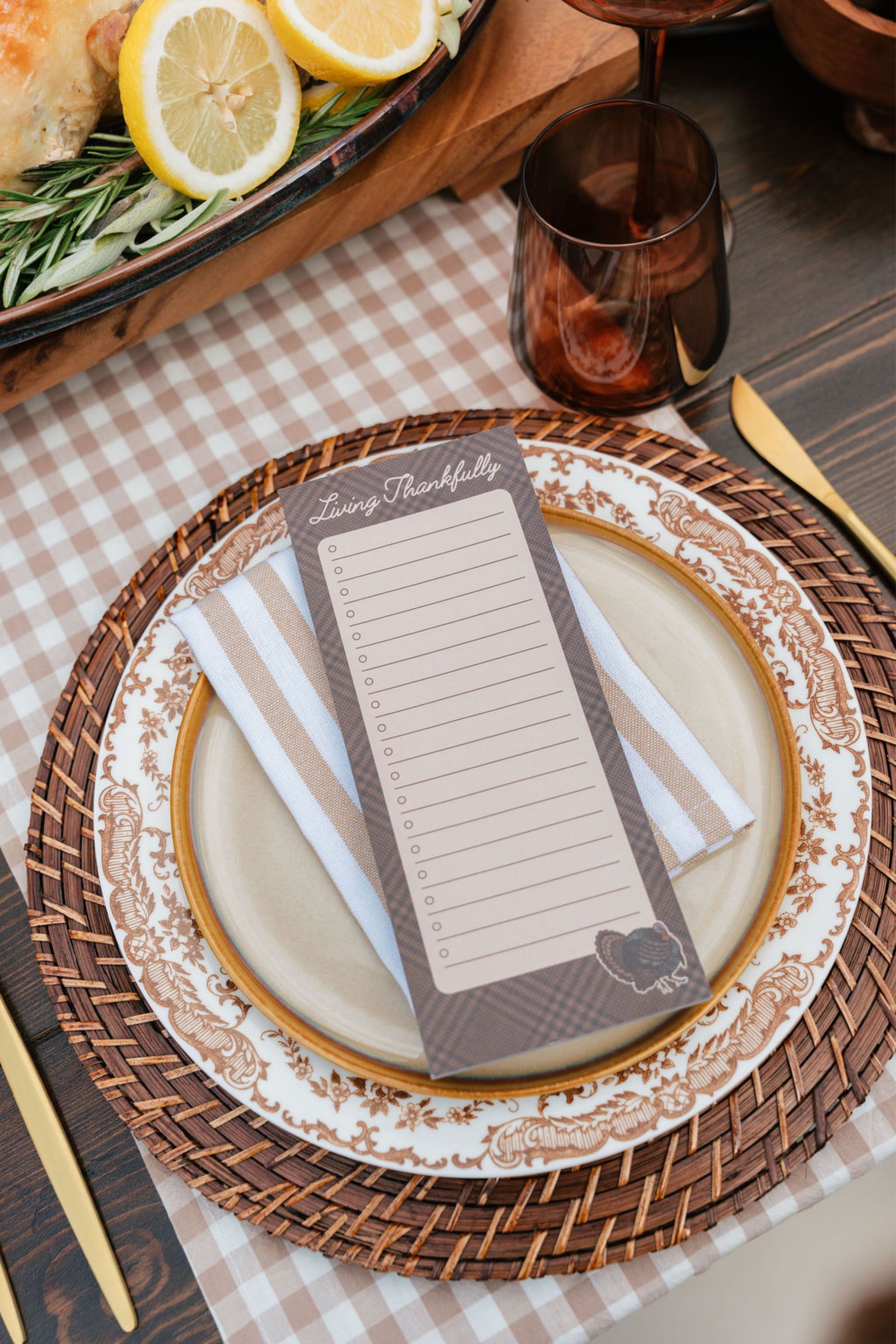 A close-up of a styled table setting featuring a rectangular notepad with lined writing space and the heading 'Living thankfully' placed on top of a place setting. The notepad is layered over a striped napkin, a decorative plate, a woven placemat, and a checkered tablecloth, with a glass, lemon slices, and a wooden serving board in the background.