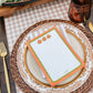 A styled tabletop place setting with a white notepad featuring orange and green borders and small pumpkin illustrations, layered on patterned plates and a woven placemat. A gingham placemat, gold-toned cutlery, amber drink glass, and a decorative tray with lemons and rosemary are also visible.