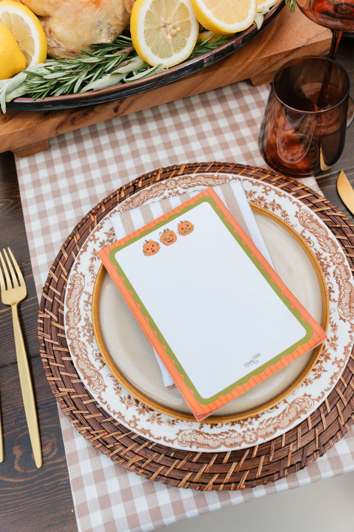 A styled tabletop place setting with a white notepad featuring orange and green borders and small pumpkin illustrations, layered on patterned plates and a woven placemat. A gingham placemat, gold-toned cutlery, amber drink glass, and a decorative tray with lemons and rosemary are also visible.