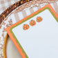 A close-up of a Halloween-themed notepad with three cute orange pumpkin illustrations, styled on a patterned plate and checkered tablecloth.