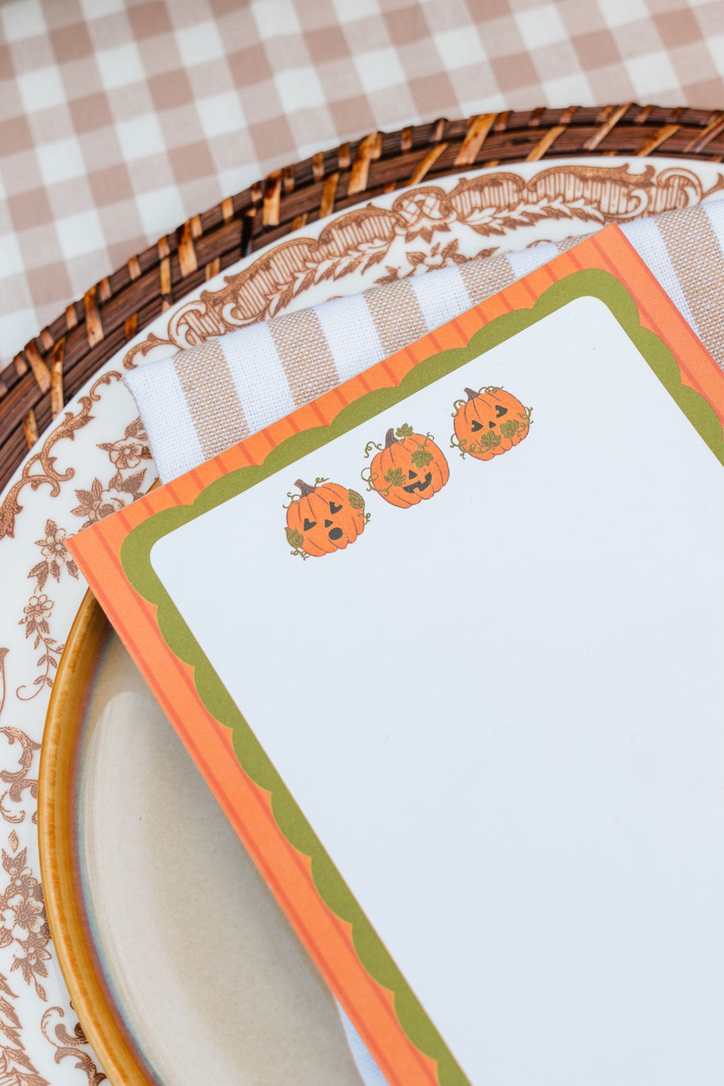 A close-up of a Halloween-themed notepad with three cute orange pumpkin illustrations, styled on a patterned plate and checkered tablecloth.