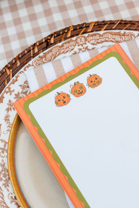 A close-up of a Halloween-themed notepad with three cute orange pumpkin illustrations, styled on a patterned plate and checkered tablecloth.