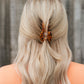 A blonde woman viewed from the back, wearing a tortoiseshell hair claw clip to secure a half-up style, with a blurred wooden fence in the background.