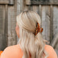 A blonde woman seen from behind wearing a brown and black leopard-print hair claw clip, set against a rustic wooden fence background.