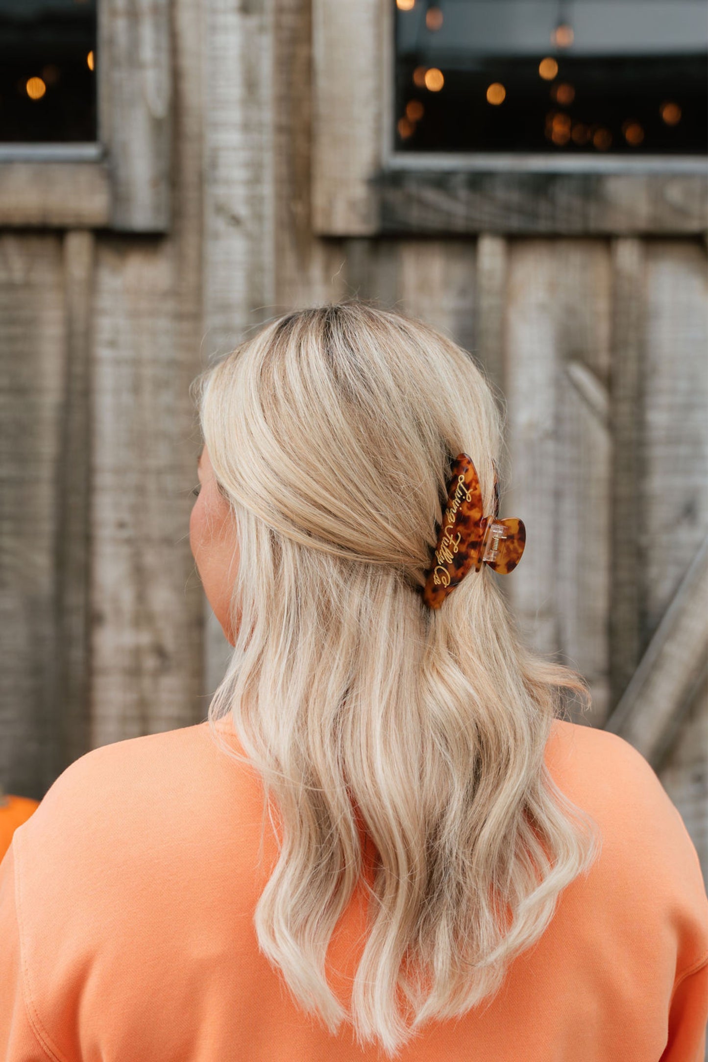 A blonde woman seen from behind wearing a brown and black leopard-print hair claw clip, set against a rustic wooden fence background.