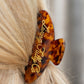 A close-up of a blonde person's hair secured with a tortoiseshell hair clip featuring gold cursive script that reads 'Living fully Co.'.