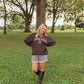 A woman stands on a grassy park lawn wearing a dark brown sweatshirt with 'Living Faith 60' printed in white, gray shorts, and dark brown knee-high boots.