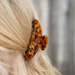 A close-up shot of a blonde person wearing a tortoiseshell hair claw clip with gold cursive lettering that reads 'Living fully Co.' against a blurred gray background.