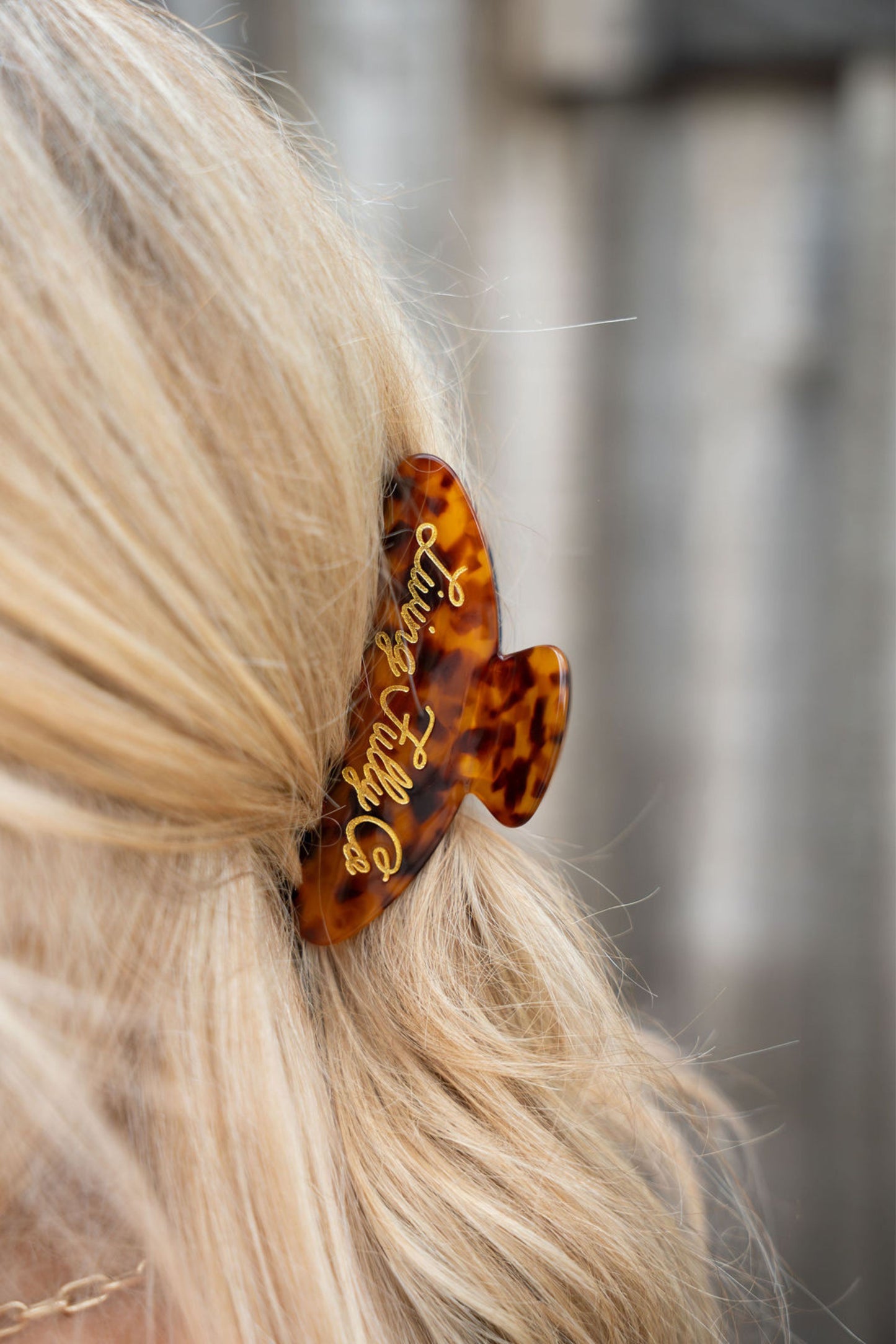 A close-up shot of a blonde person wearing a tortoiseshell hair claw clip with gold cursive lettering that reads 'Living fully Co.' against a blurred gray background.