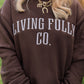 A woman wearing a dark brown crewneck sweatshirt with 'LIVING FULLY CO.' printed in white arched lettering across the chest, photographed outdoors with a blurred green field in the background.