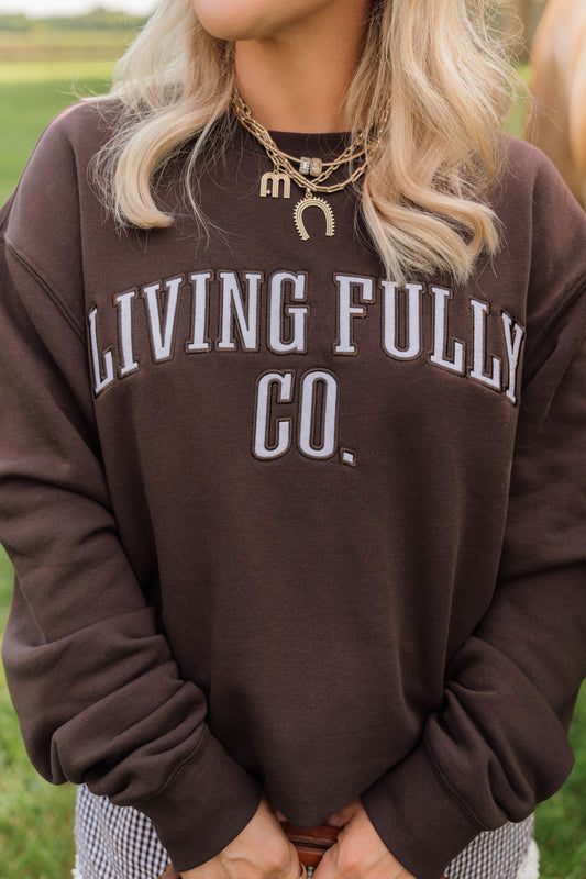 A woman wearing a dark brown crewneck sweatshirt with 'LIVING FULLY CO.' printed in white arched lettering across the chest, photographed outdoors with a blurred green field in the background.