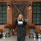 A woman stands on a porch in front of a wooden barn-style door, wearing a black long-sleeve top with a white graphic and a black skirt with white polka dots. She holds a checkered bowl, with potted orange flowers and black-and-white checkered planters on either side.