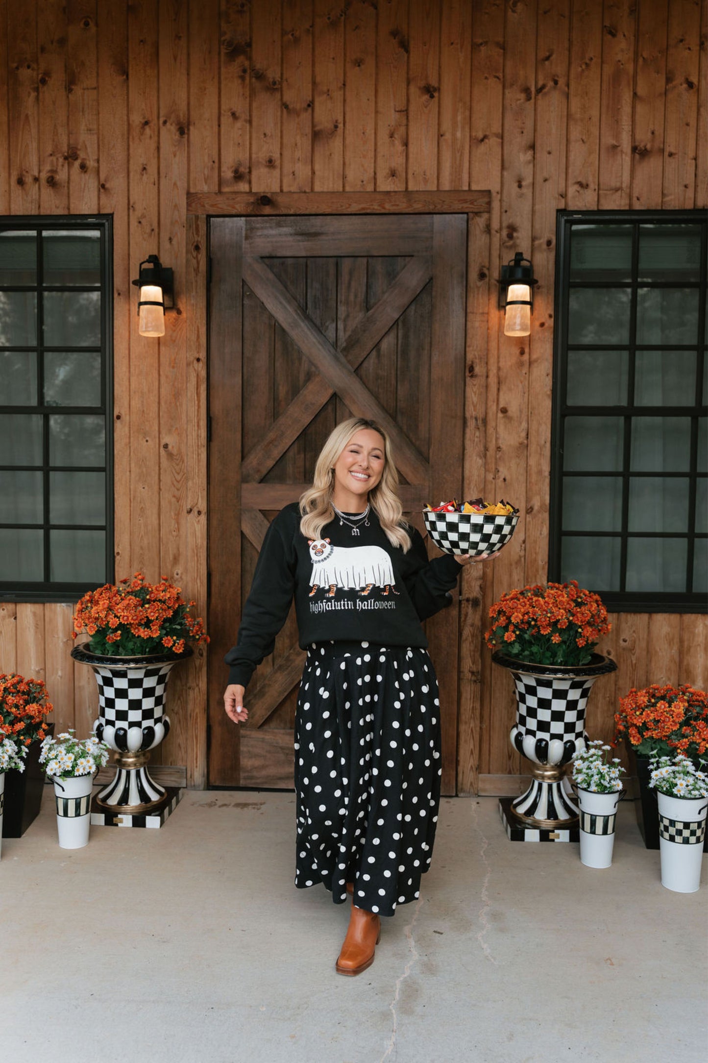 A woman stands on a porch in front of a wooden barn-style door, wearing a black long-sleeve top with a white graphic and a black skirt with white polka dots. She holds a checkered bowl, with potted orange flowers and black-and-white checkered planters on either side.