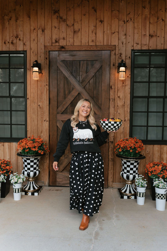 A woman stands on a porch in front of a wooden barn-style door, wearing a black long-sleeve top with a white graphic and a black skirt with white polka dots. She holds a checkered bowl, with potted orange flowers and black-and-white checkered planters on either side.