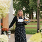 A woman stands under a wooden porch wearing a black long-sleeve top with a white graphic and a long black skirt with white polka dots. She is holding a checkered bowl filled with fruit, with flowers and a garden visible in the background.