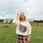 A woman stands in a grassy field with farm buildings and a cloudy sky in the background, wearing a cream sweatshirt with a large turkey graphic and a brown plaid skirt.