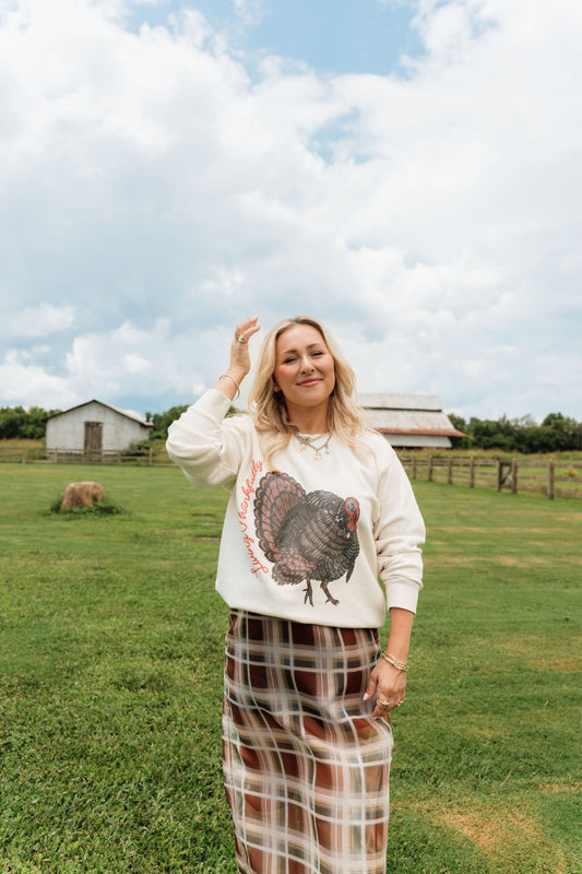 A woman stands in a grassy field with farm buildings and a cloudy sky in the background, wearing a cream sweatshirt with a large turkey graphic and a brown plaid skirt.