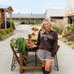A woman stands outdoors on a farm path wearing a dark brown sweatshirt with 'LIVING FULLY CO.' printed in white across the chest, paired with gray shorts and dark brown knee-high boots.