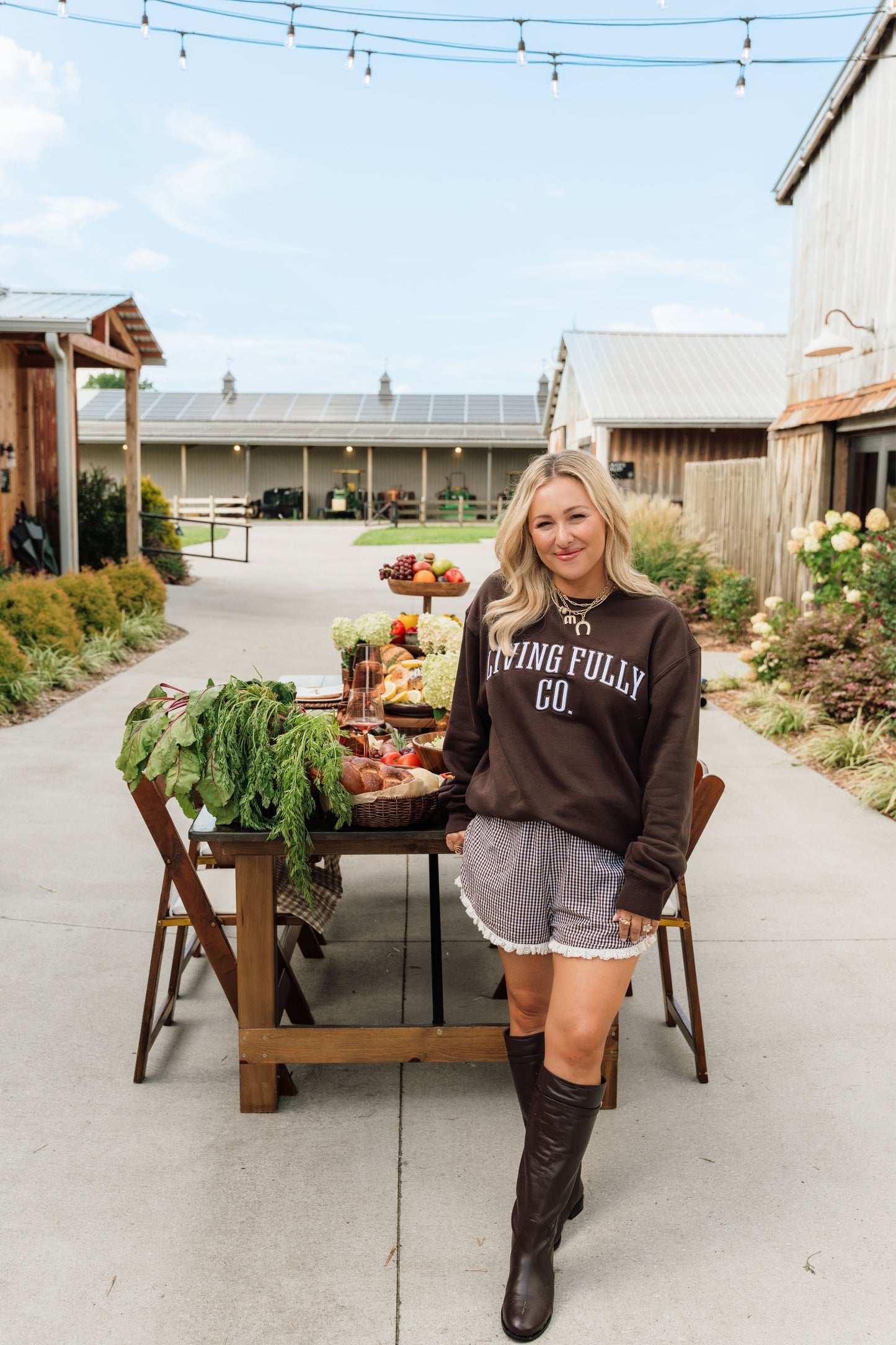 A woman stands outdoors on a farm path wearing a dark brown sweatshirt with 'LIVING FULLY CO.' printed in white across the chest, paired with gray shorts and dark brown knee-high boots.