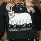 A woman wearing a black crewneck sweatshirt with a white ghost graphic and the text 'highfalutin halloween' in a gothic font. The sweatshirt also features a small cartoon animal wearing a skull mask and a black chain necklace.