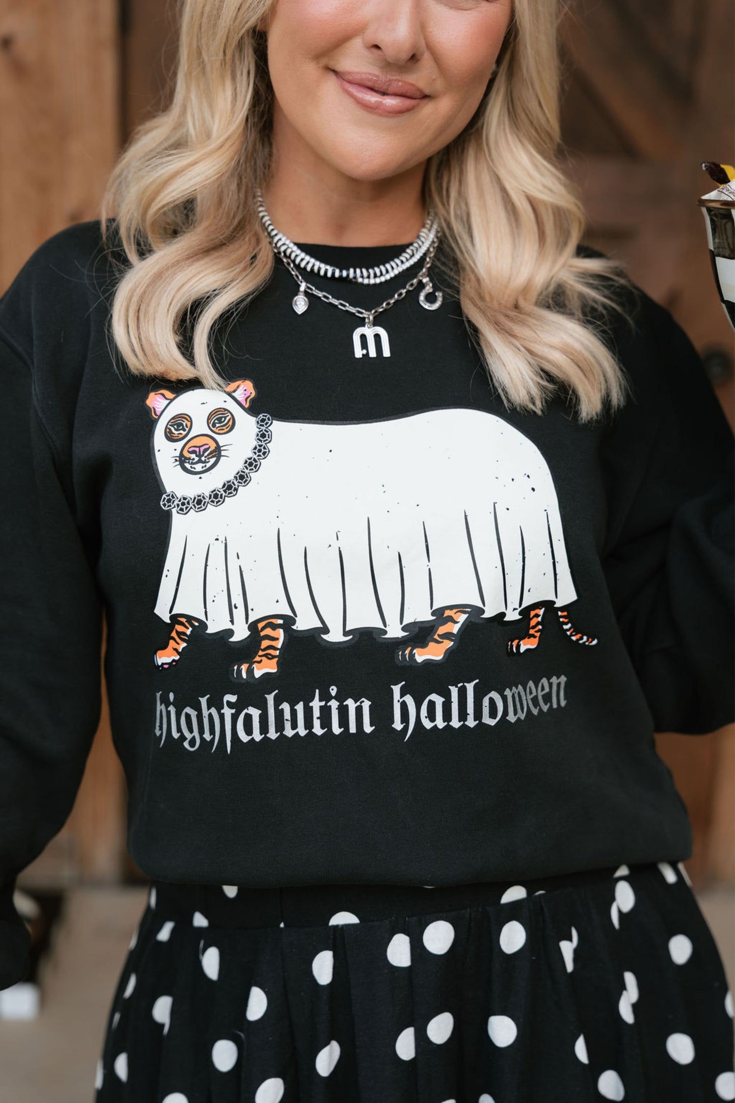 A woman wearing a black crewneck sweatshirt with a white ghost graphic and the text 'highfalutin halloween' in a gothic font. The sweatshirt also features a small cartoon animal wearing a skull mask and a black chain necklace.