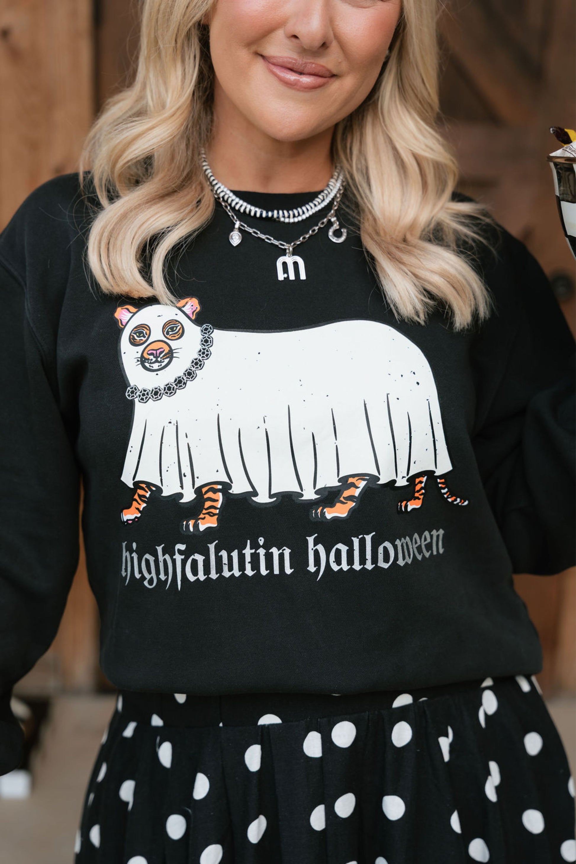 A woman wearing a black crewneck sweatshirt with a white ghost graphic and the text 'highfalutin halloween' in a gothic font. The sweatshirt also features a small cartoon animal wearing a skull mask and a black chain necklace.