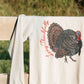 An off-white long-sleeve sweatshirt draped over a wooden fence outdoors, featuring a graphic print of a brown and black turkey with red script text that reads 'Thankfully Living'.