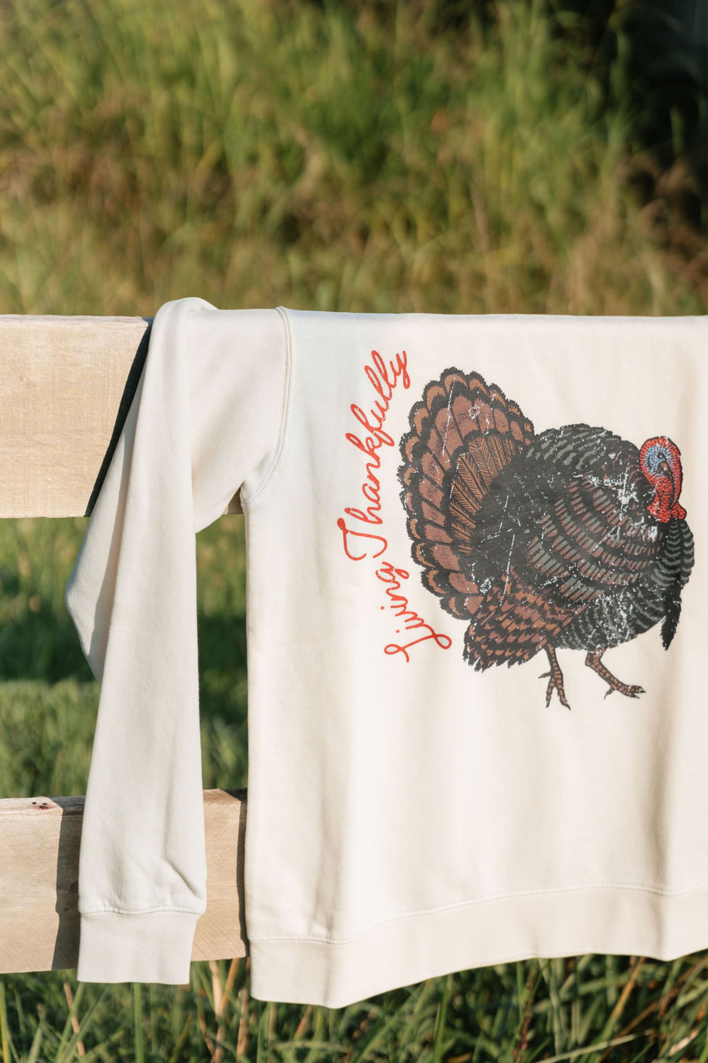 An off-white long-sleeve sweatshirt draped over a wooden fence outdoors, featuring a graphic print of a brown and black turkey with red script text that reads 'Thankfully Living'.