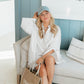 Woman sitting on a couch wearing a white shirt and blue cap, holding a brown tote bag with 'Living fully' text.