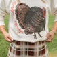 A woman outdoors wearing a cream-colored sweatshirt with a large distressed black and brown turkey graphic and red cursive text, paired with a brown and beige plaid skirt.