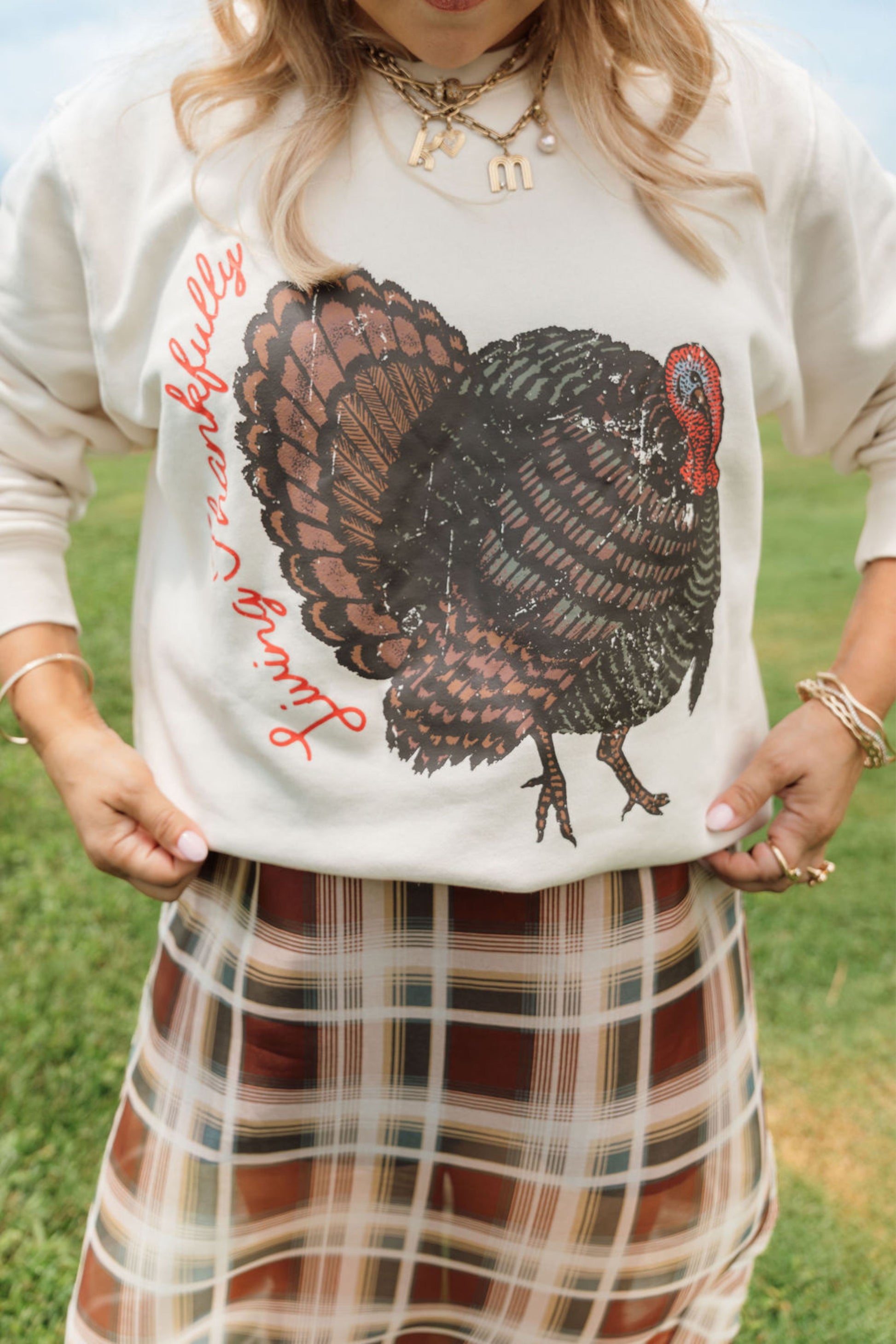 A woman outdoors wearing a cream-colored sweatshirt with a large distressed black and brown turkey graphic and red cursive text, paired with a brown and beige plaid skirt.