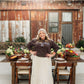 A woman stands outdoors in front of a rustic wooden building, wearing a dark brown long-sleeve sweatshirt with white text that reads 'LIVING FULLY CO.' and a long cream skirt with small dark polka dots.