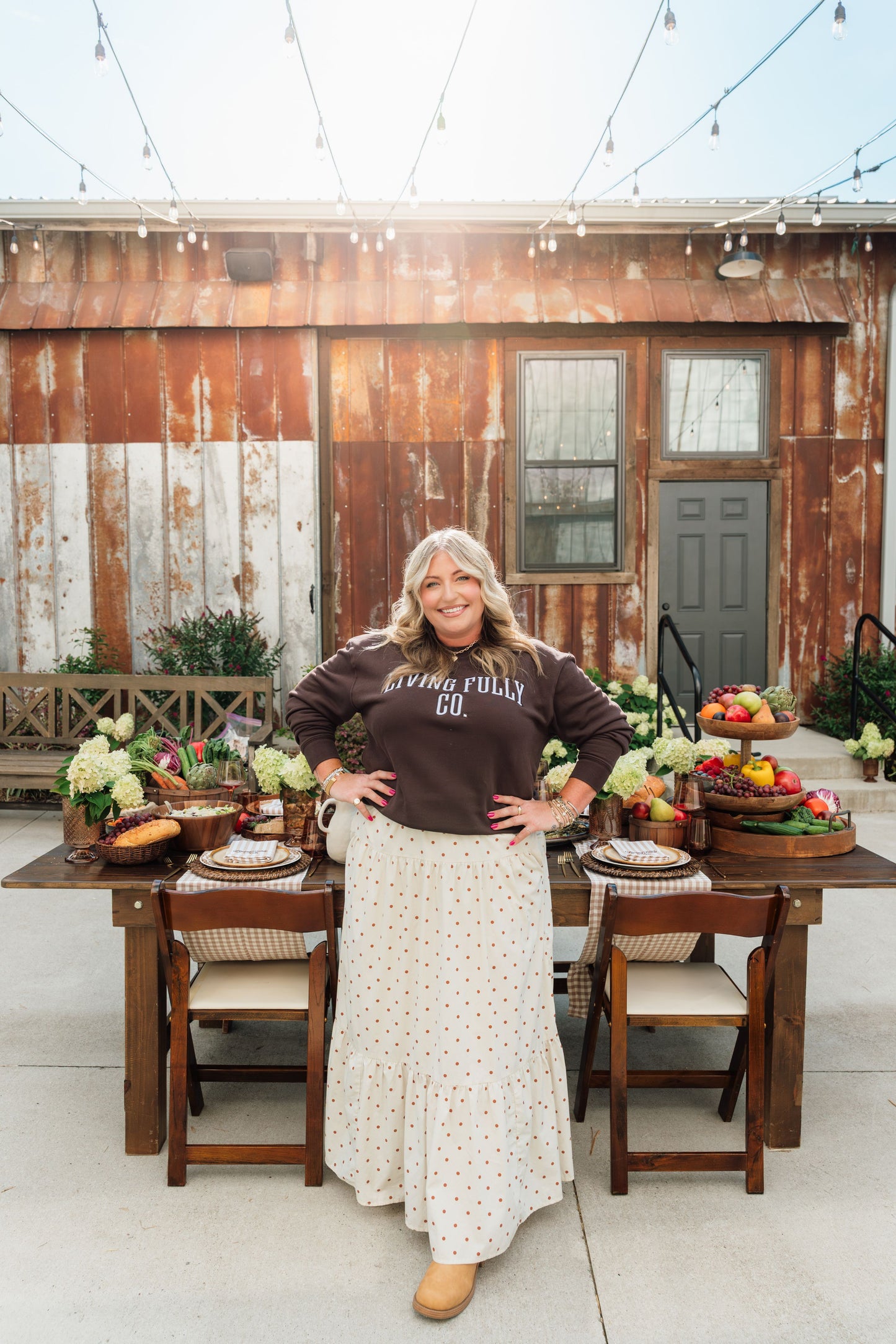 A woman stands outdoors in front of a rustic wooden building, wearing a dark brown long-sleeve sweatshirt with white text that reads 'LIVING FULLY CO.' and a long cream skirt with small dark polka dots.