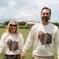 A man and a woman holding hands, standing in a grassy field under a cloudy sky. Both are wearing matching cream-colored crewneck sweatshirts featuring a large graphic of a brown and black turkey with red accents.