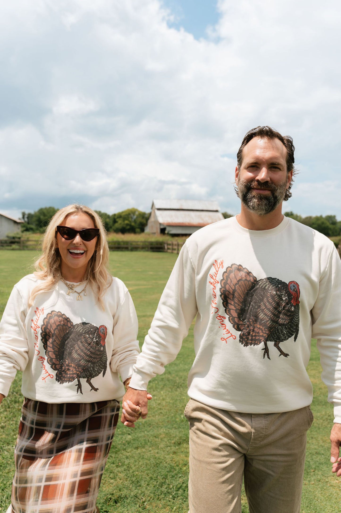 A man and a woman holding hands, standing in a grassy field under a cloudy sky. Both are wearing matching cream-colored crewneck sweatshirts featuring a large graphic of a brown and black turkey with red accents.