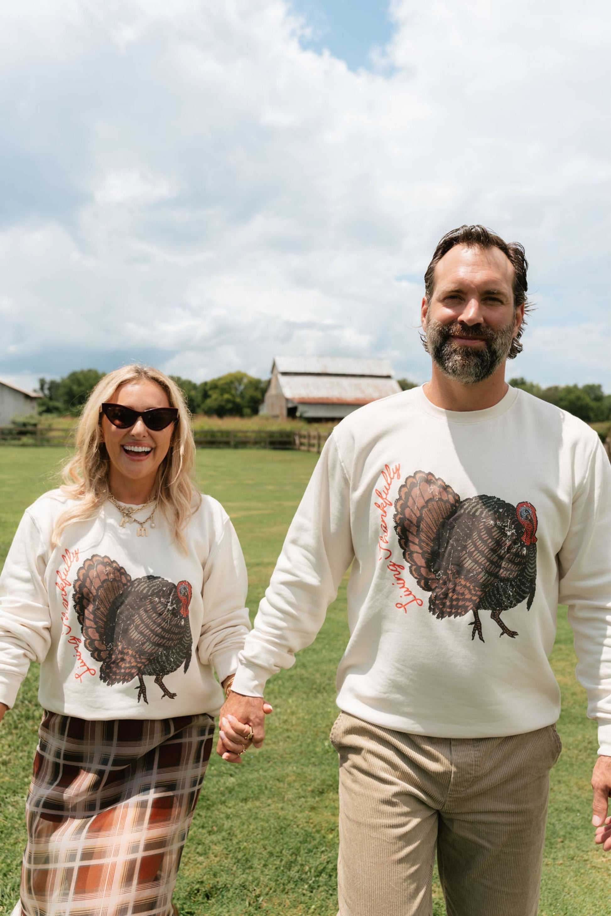 A man and a woman holding hands, standing in a grassy field under a cloudy sky. Both are wearing matching cream-colored crewneck sweatshirts featuring a large graphic of a brown and black turkey with red accents.