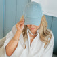 Woman wearing a light blue cap, white shirt, and pearl necklace against a light blue wall.
