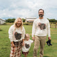 A family of four stands on a grassy farm field under a cloudy sky, wearing matching cream-colored long-sleeve sweatshirts with a large black and brown turkey graphic on the front. The woman wears a brown and beige plaid skirt, the man wears khaki pants and holds a black shoe, and the child wears a plaid skirt.