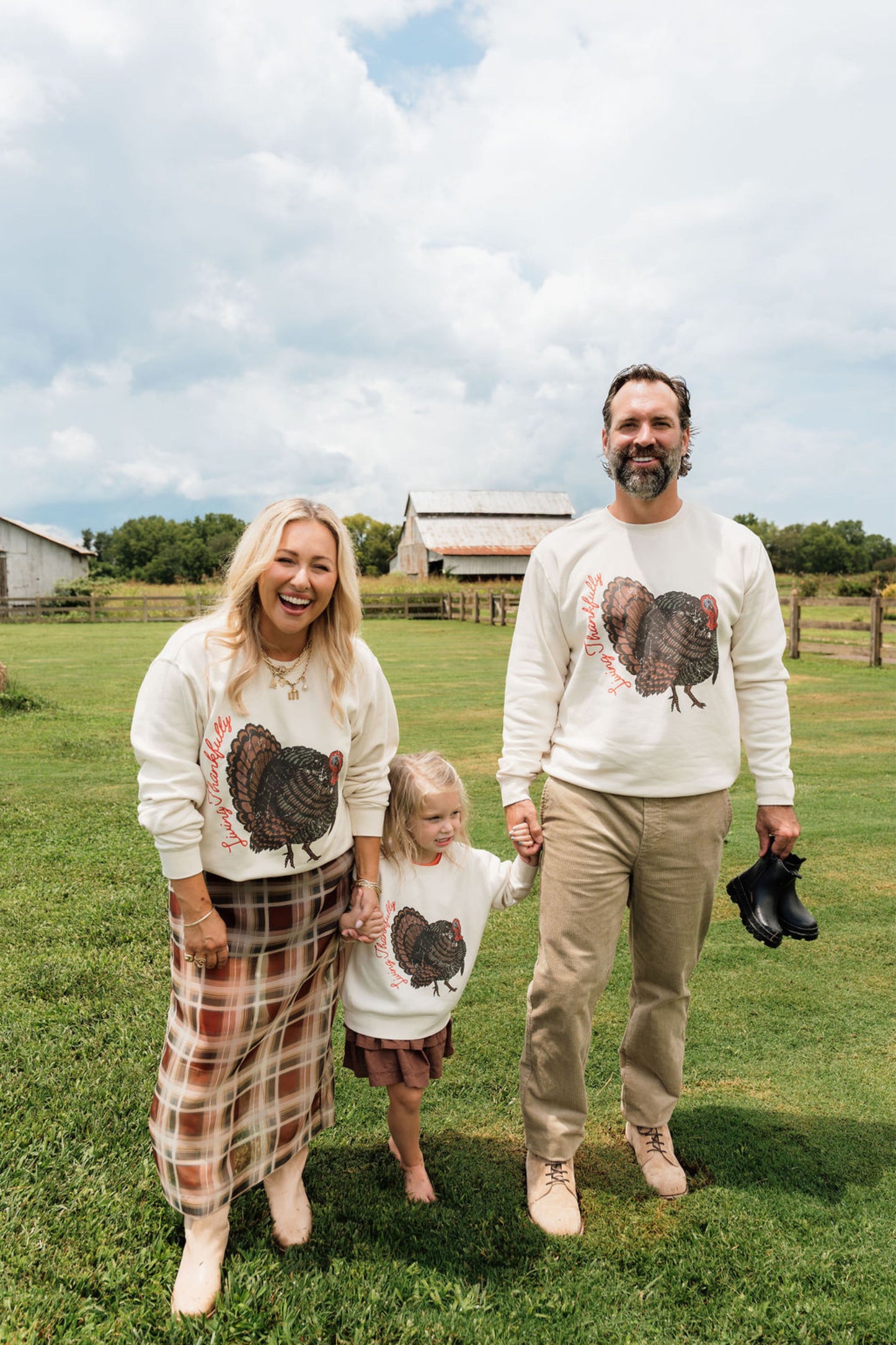 A family of four stands on a grassy farm field under a cloudy sky, wearing matching cream-colored long-sleeve sweatshirts with a large black and brown turkey graphic on the front. The woman wears a brown and beige plaid skirt, the man wears khaki pants and holds a black shoe, and the child wears a plaid skirt.