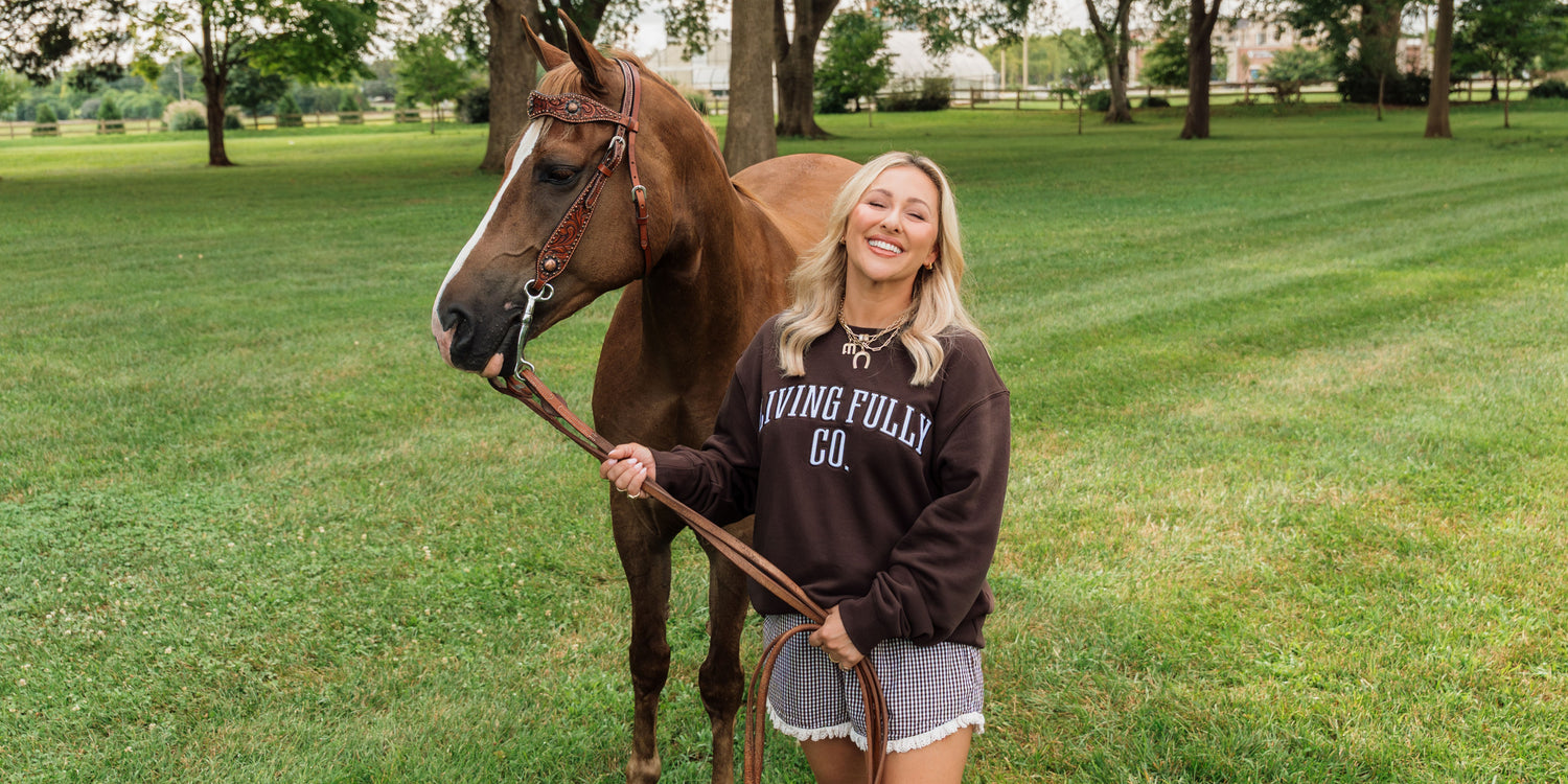 A woman stands in a grassy park next to a brown horse, wearing a dark brown sweatshirt with 'LIVING FULLY CO.' printed in white across the chest.