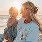 Woman and child on a beach at sunset, wearing a light blue sweatshirt with a festive, Independence Day design.