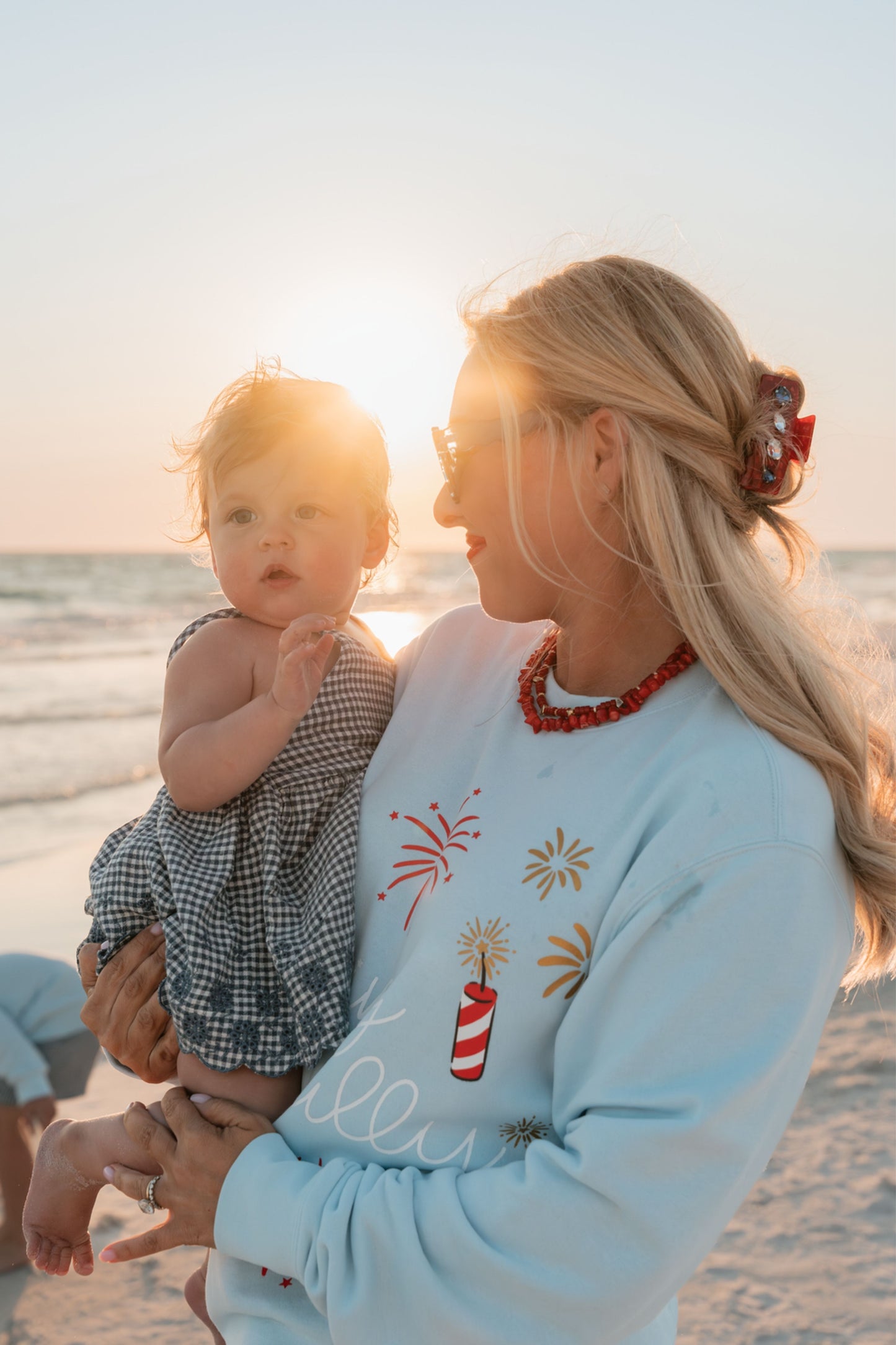 Woman and child on a beach at sunset, wearing a light blue sweatshirt with a festive, Independence Day design.