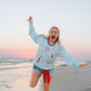 Person in a light blue LFC sweatshirt and denim shorts standing on a beach with a colorful sky.