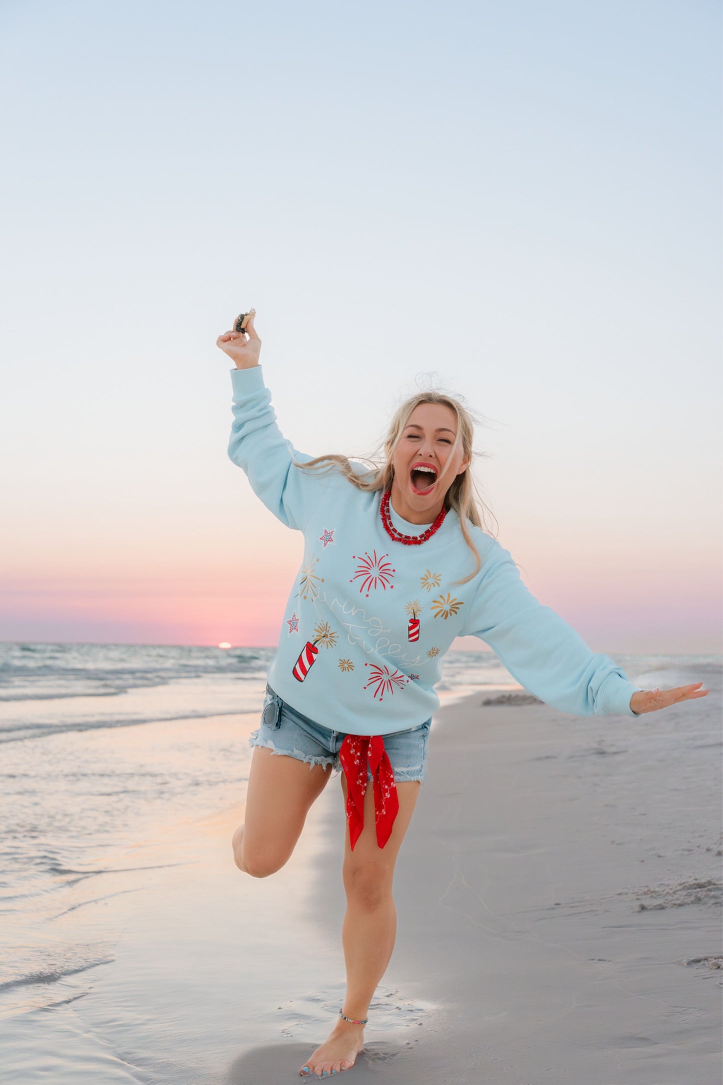 Person in a light blue LFC sweatshirt and denim shorts standing on a beach with a colorful sky.