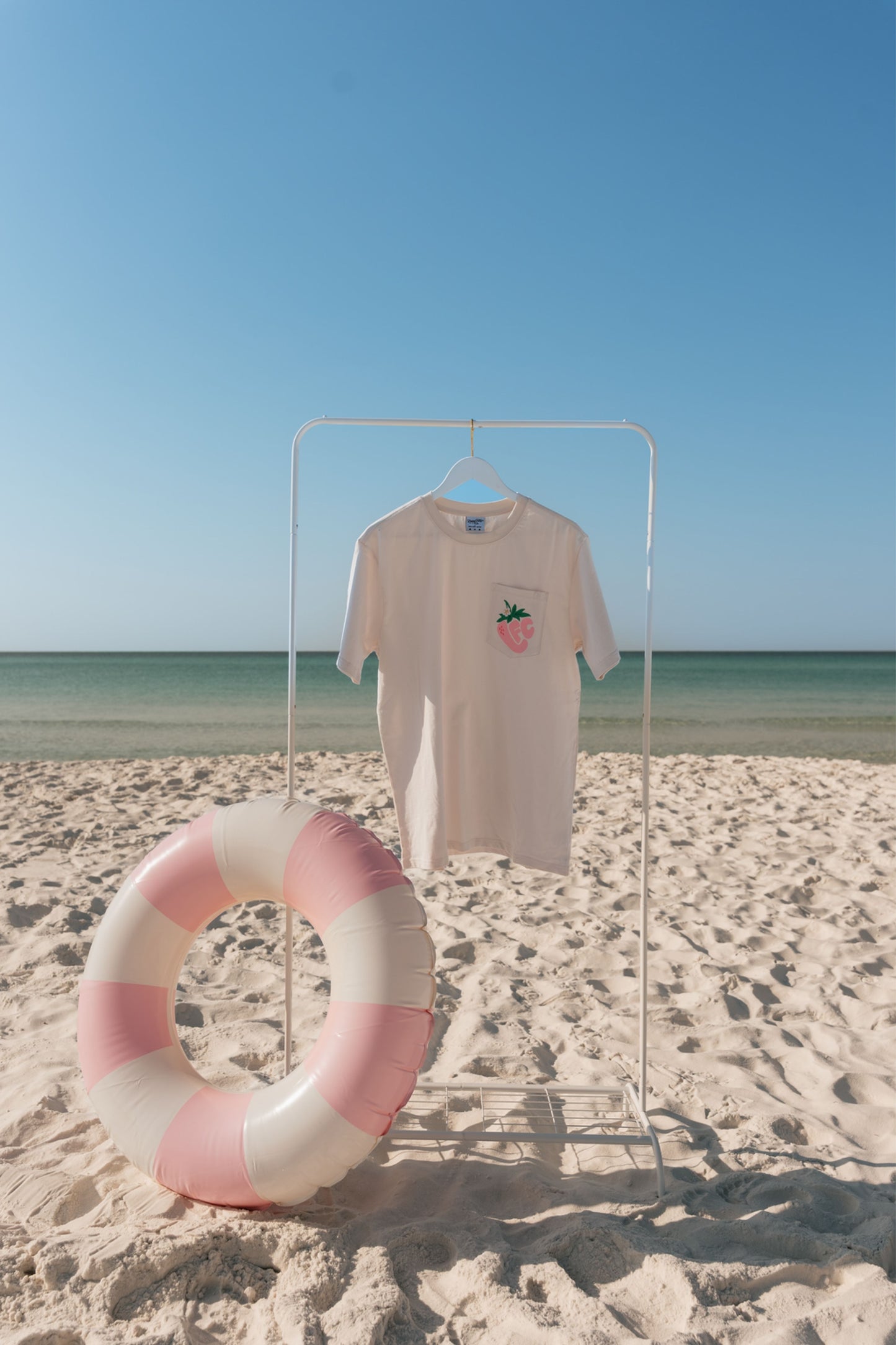T-shirt with a strawberry design on a stand next to a life buoy on a sandy beach.
