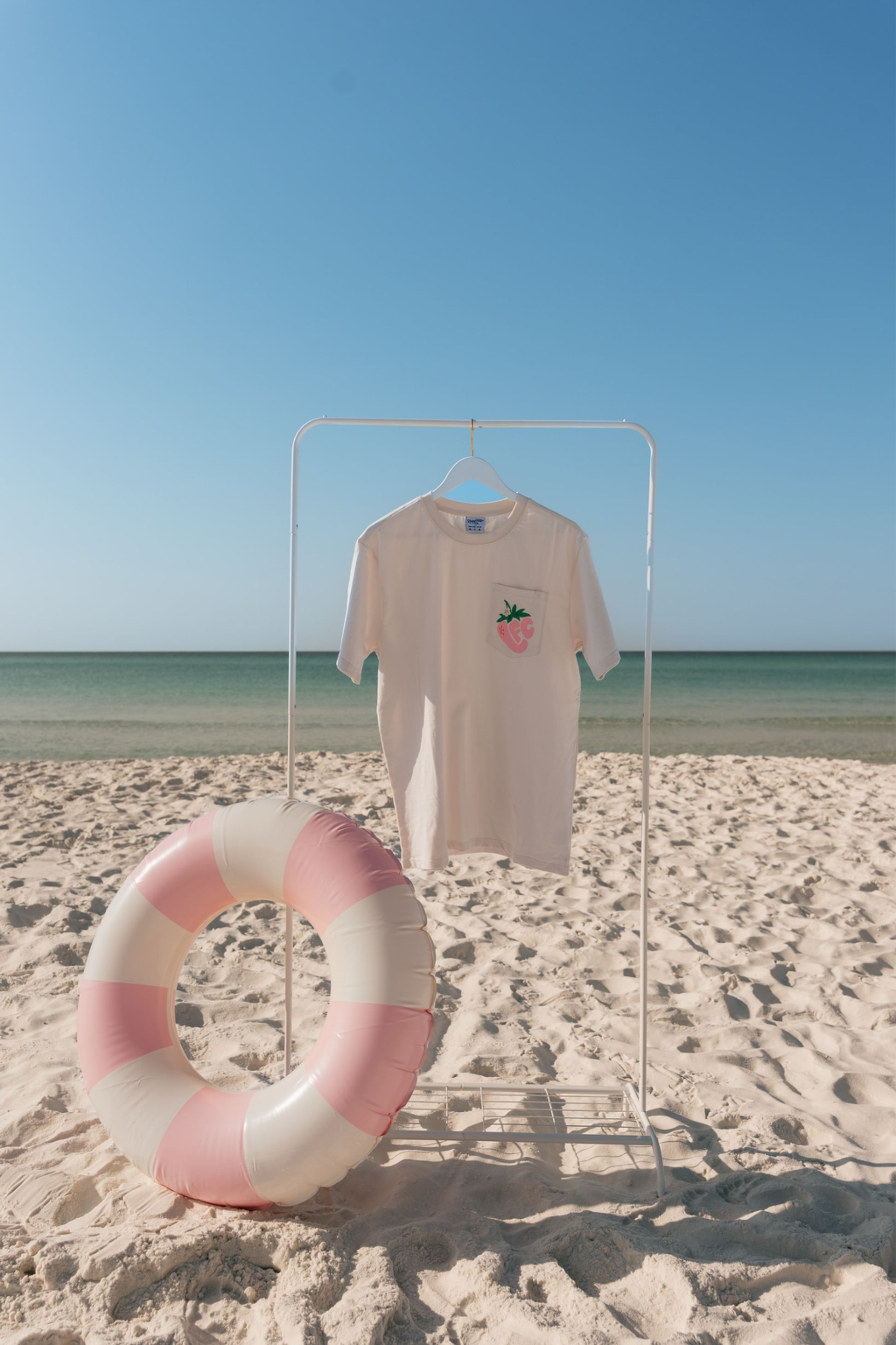 T-shirt with a strawberry design on a stand next to a life buoy on a sandy beach.
