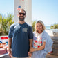 Man and woman standing outdoors with American flags and a barbecue in the background.