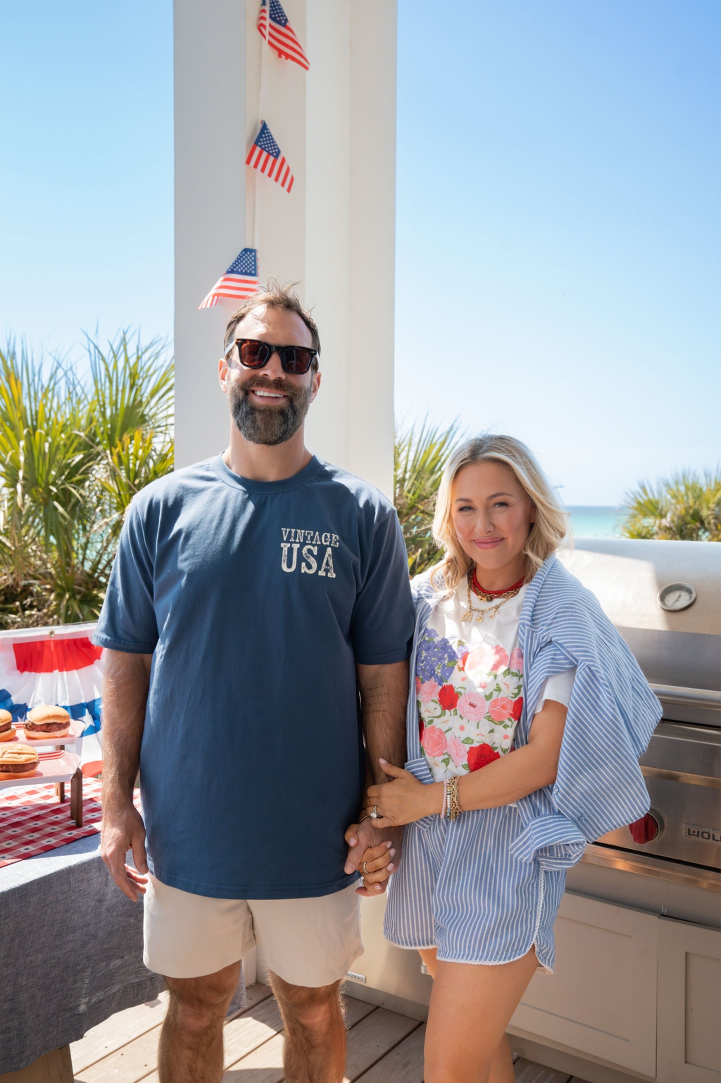 Man and woman standing outdoors with American flags and a barbecue in the background.